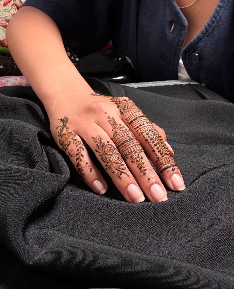 A hand with intricate NooniDesigns henna across the fingers and knuckles on dark fabric.